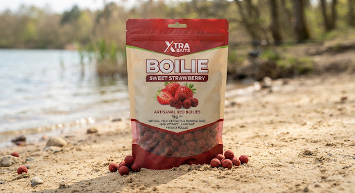 Artisanal red boilies 1kg pack titled XTRA Boilie Sweet Strawberry with natural fruit extracts and fishmeal base photographed on clean sand.