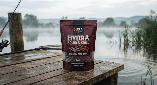 A premium 1kg bag of dark burgundy carp fishing boilies labeled Hydra Squid and Krill, set on an authentic natural wooden fishing pier by the serene misty morning lake, soft atmospheric diffused daylight, organic high-end fishing product photography.