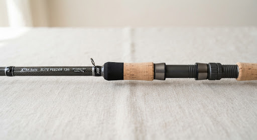 Studio product photograph of a modern high modulus carbon fiber feeder fishing rod handle with premium cork grip on clean linen surface