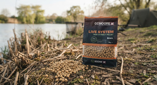 CC Moore Live System Pellets 1kg bag shown upright alongside scattered creamy light brown 6mm pellets on natural reed lake bank, soft warm lighting, refined outdoor British specimen carp fishing styling.
